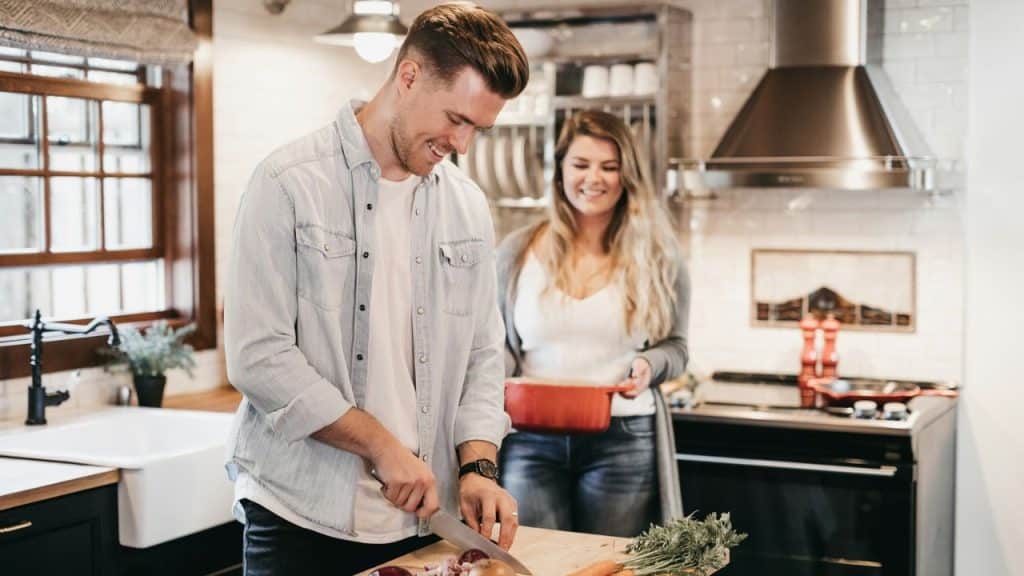 Man chopping vegetables in a kitchen while a woman smiles holding a pot.