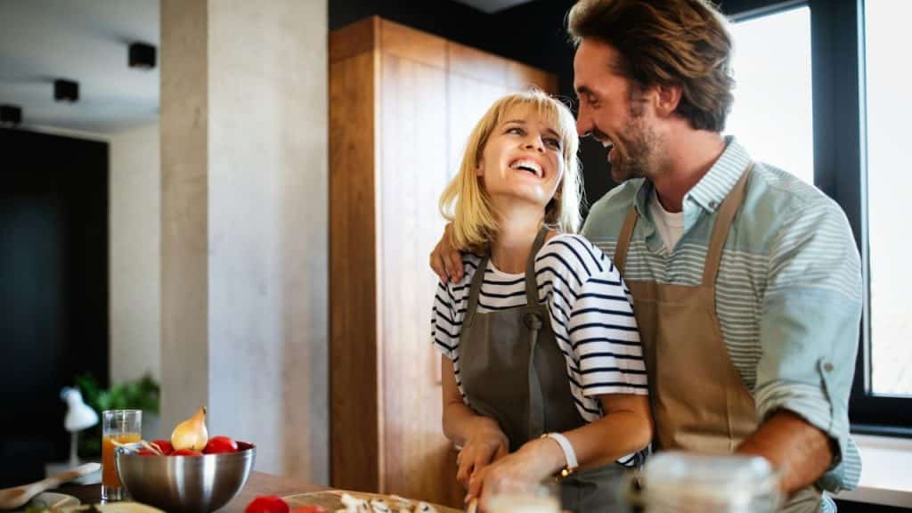 A smiling man embraces a laughing woman from behind while they cook in a kitchen.