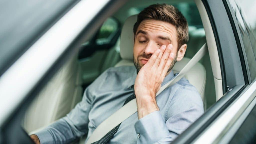 Man in a shirt and tie sitting in a car, rubbing his tired eyes.