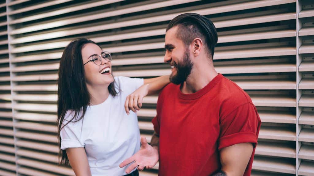 Smiling couple leaning against a slatted wall, the woman wearing glasses.