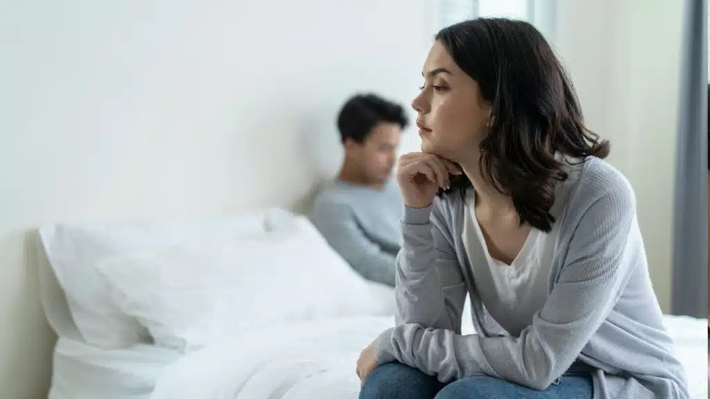 A woman looking sad and sitting on the edge of a bed while her husband lies on it in the background.