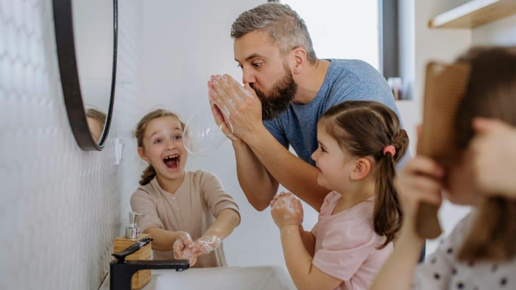 Father having fun blowing bubbles from soap in the bathroom with his little daughters.
