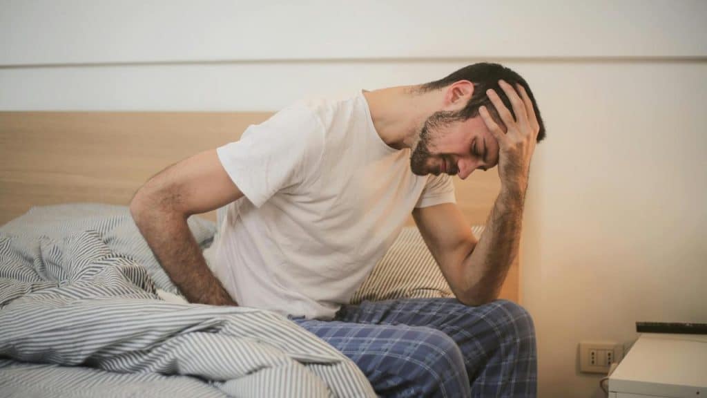 A man sitting on a bed holding his head in discomfort.