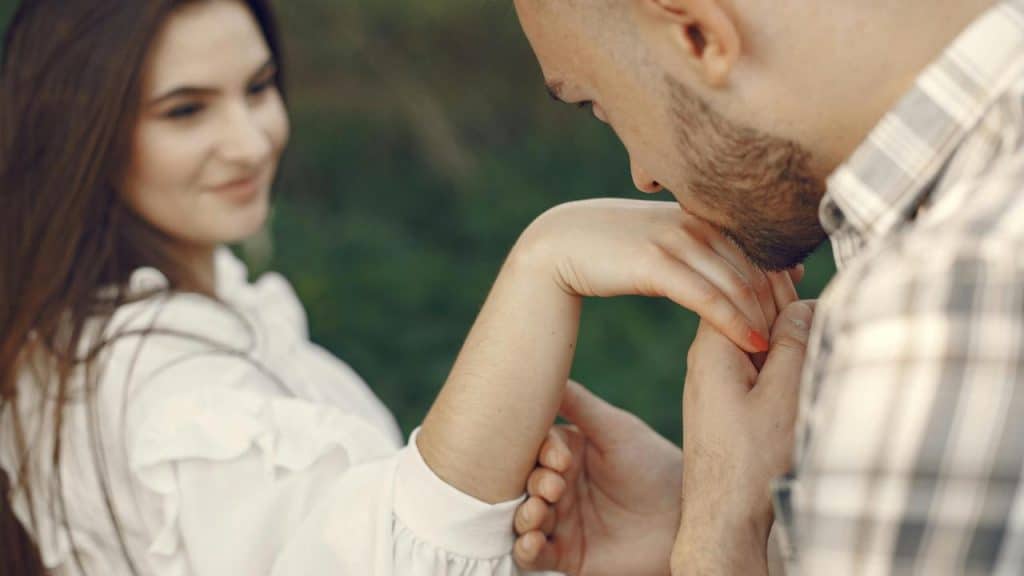 A man kissing a womanโs hand while she smiles outdoors.