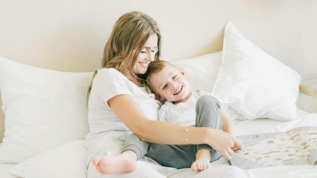 A mother and child sit on a bed smiling and cuddling together.