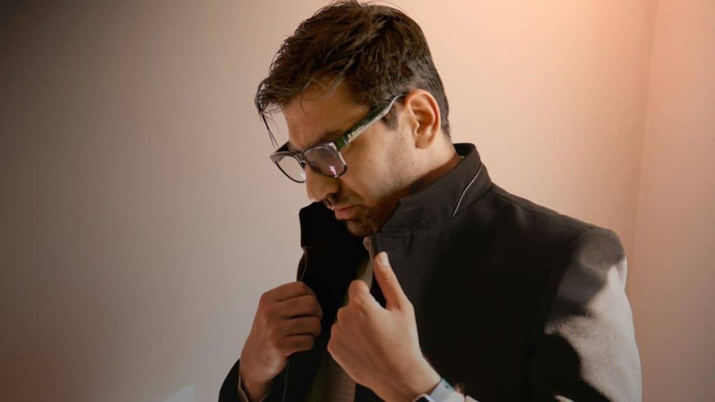 A man in glasses and a dark jacket looks down, adjusting his collar in warm light.