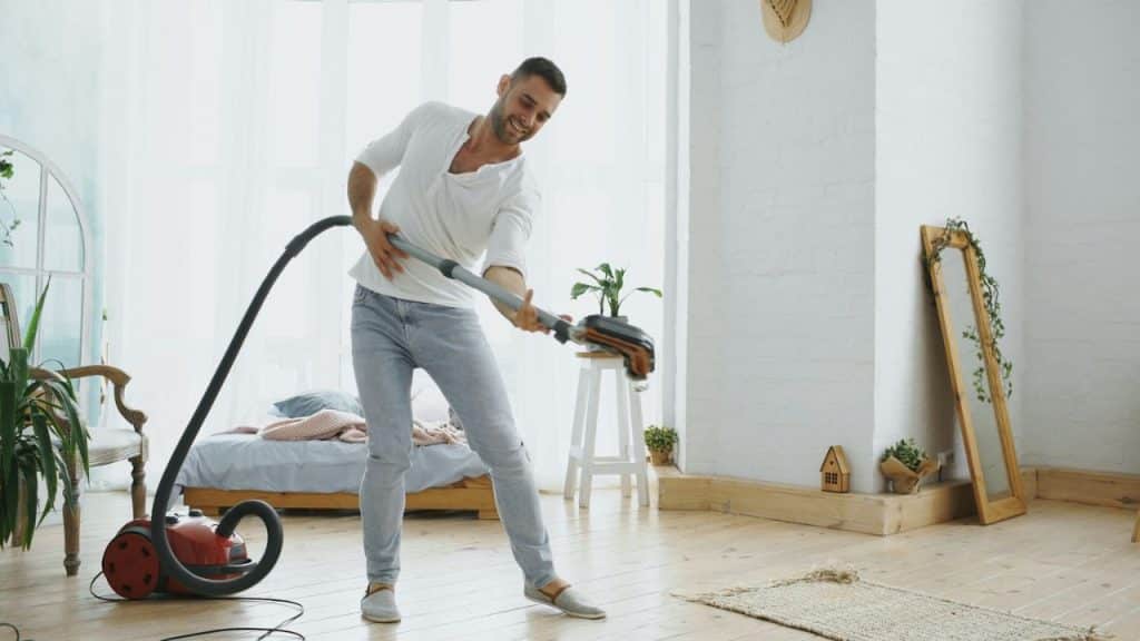 A smiling man holds a vacuum cleaner tube like a guitar while cleaning a bright room.