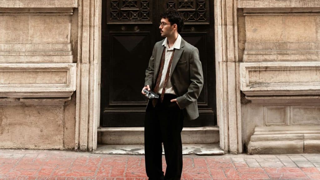 A man in a blazer and loosened tie stands outside with a camera looking away.