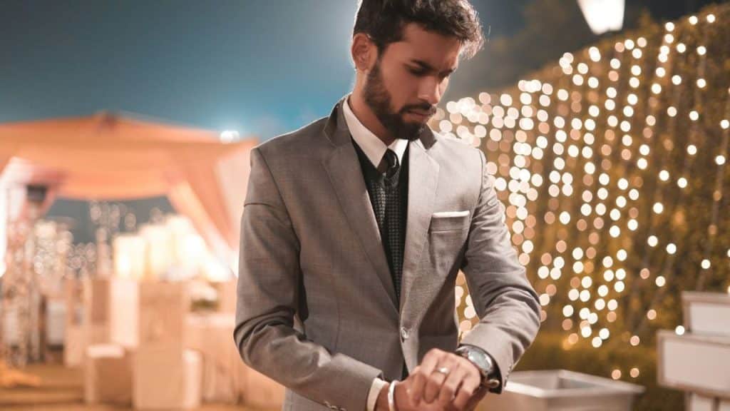 A man in a gray suit and tie looks down at his wristwatch outdoors at night.