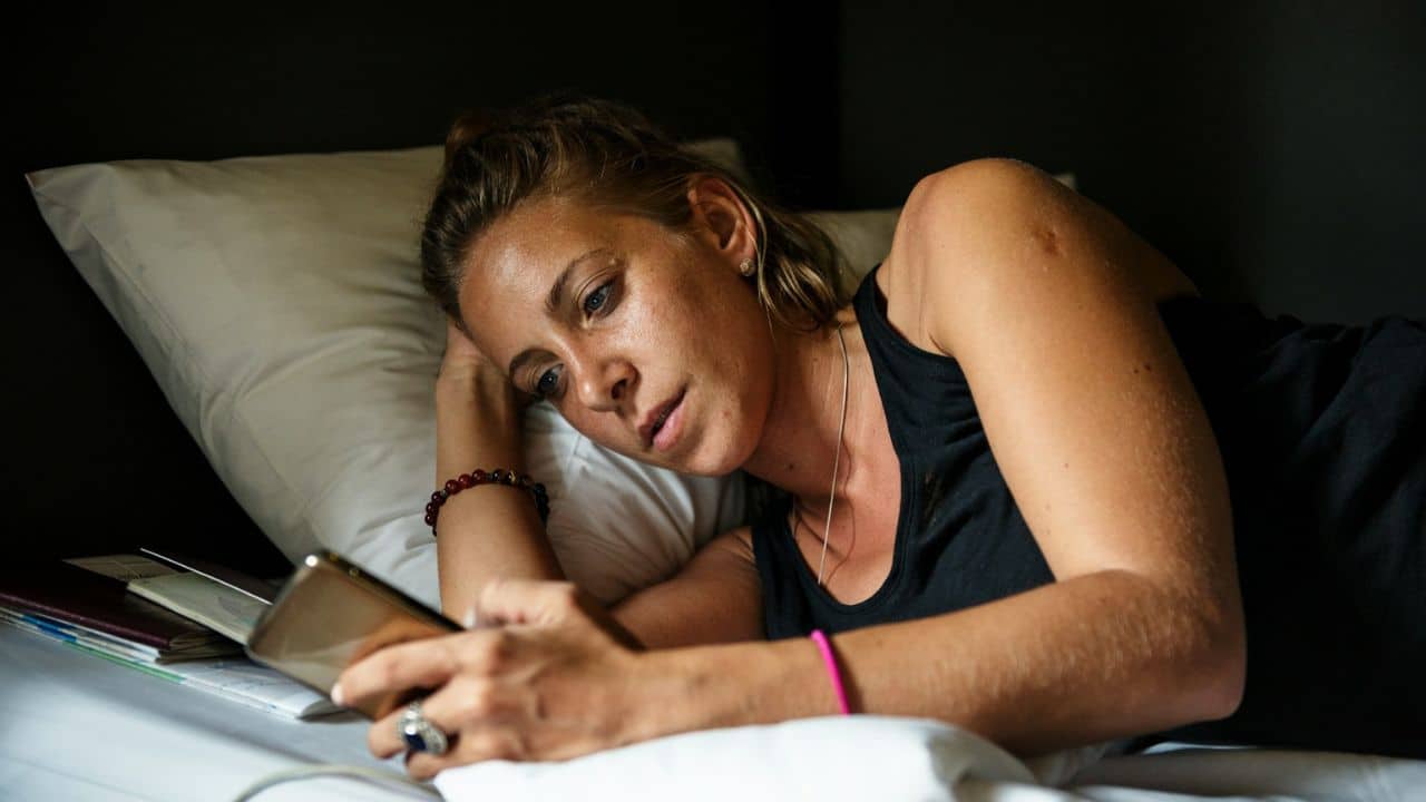 A woman with blonde hair lies on a bed, looking intently at a smartphone in her hands.