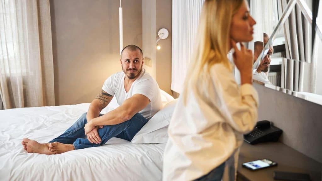 A man sits on a bed smiling while a woman stands by a mirror in a bedroom.