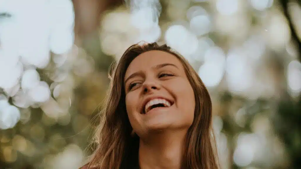 A woman laughing outdoors with soft, blurred greenery in the background.