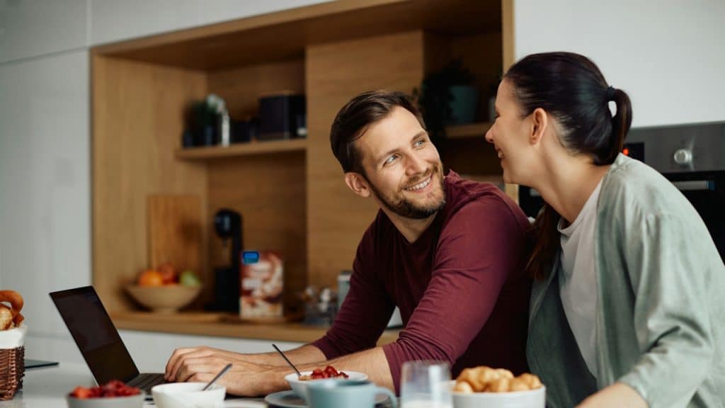 Happy couple smiling at each other while using a laptop and eating breakfast in the kitchen.