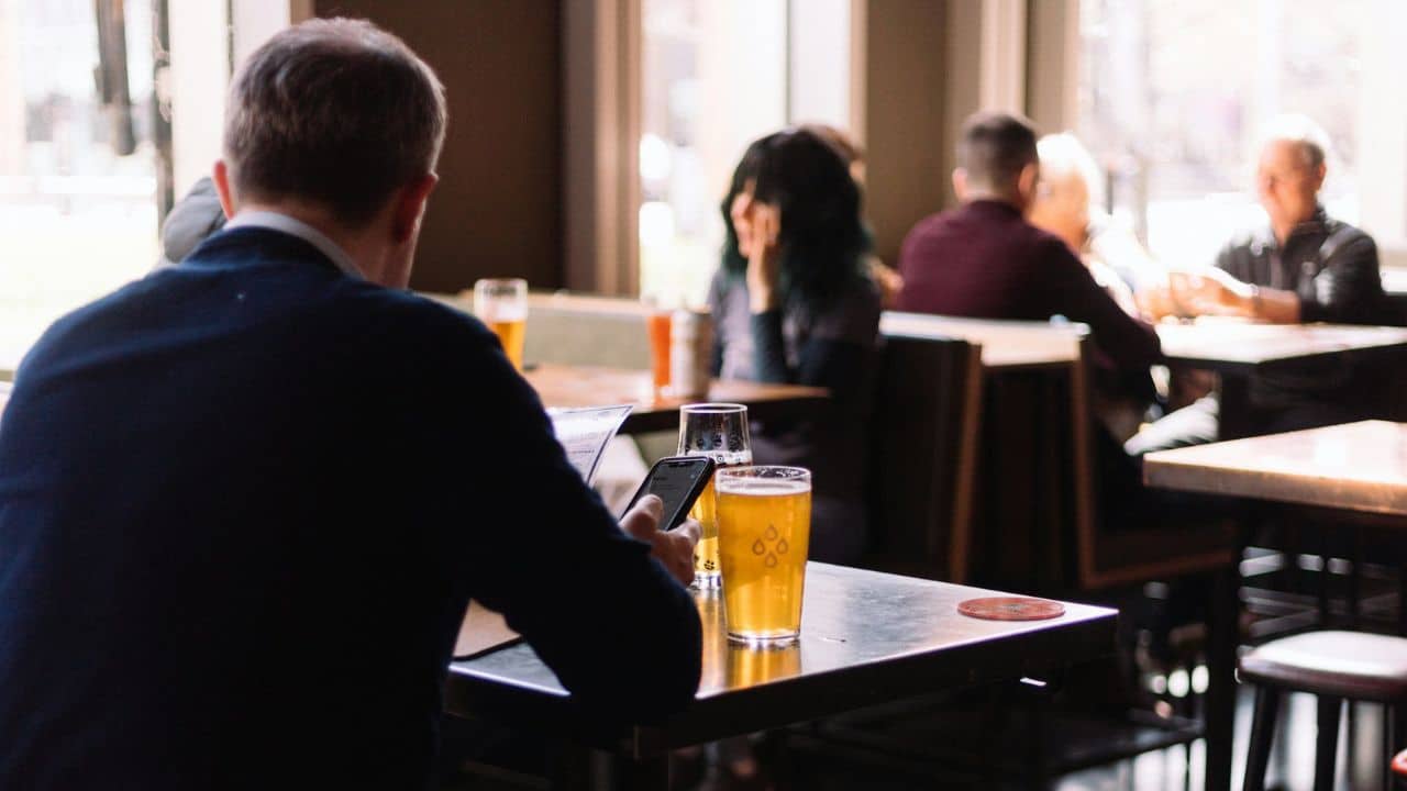 A man in a dark suit sitting at a table with two beers in a crowded, bright pub.