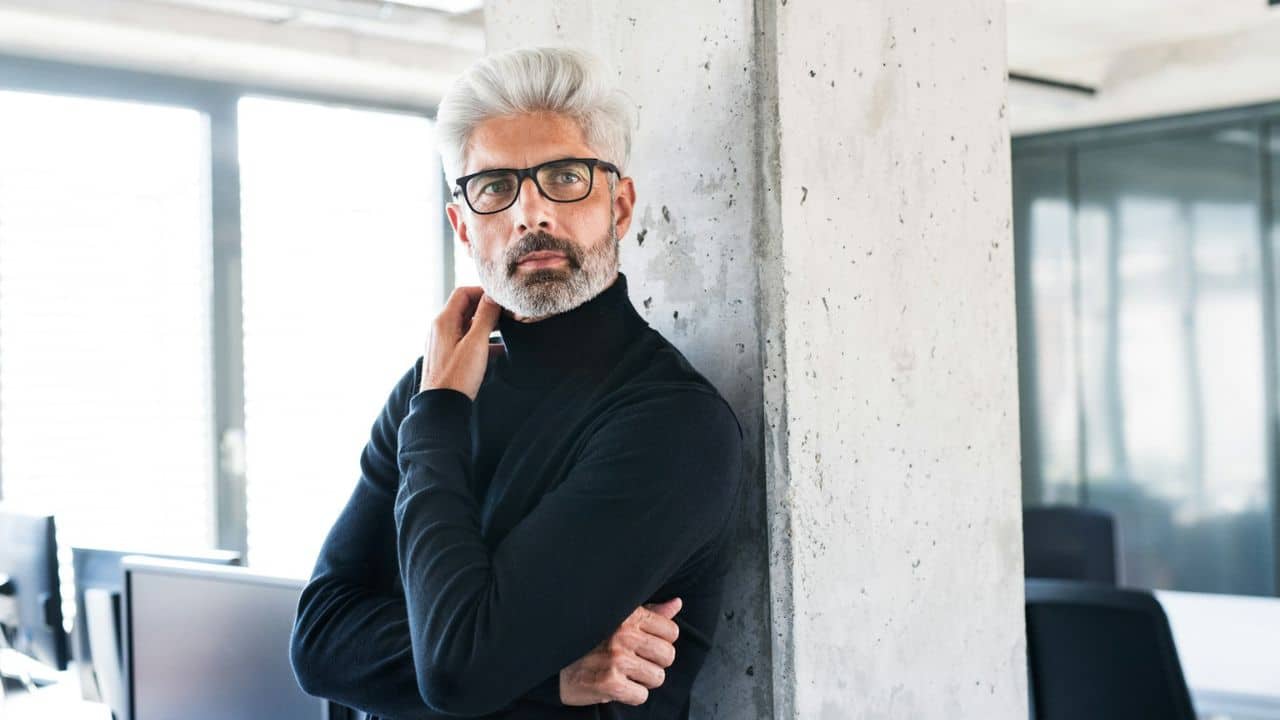 A serious man with gray hair and glasses is leaning against a concrete column indoors.