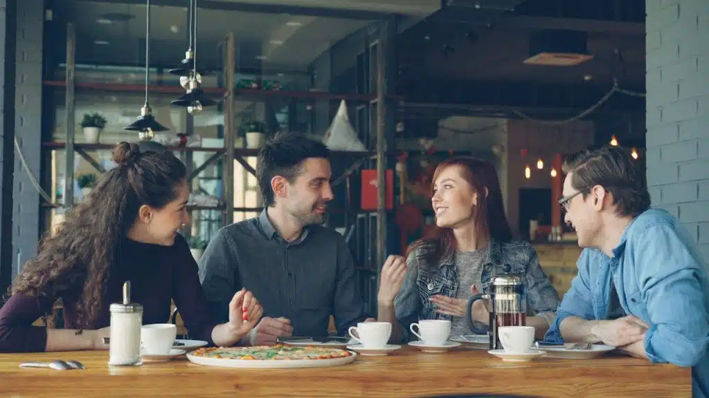 Four friends are sitting around a table in a cafe or restaurant with coffee cups and a plate of pizza, engaged in conversation.