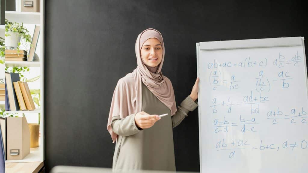 A woman wearing a hijab is teaching mathematics on a whiteboard.