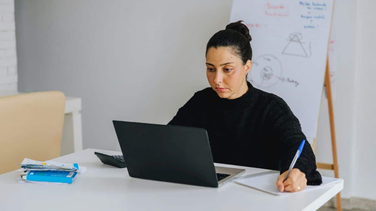 A woman is working at a white desk, looking at a laptop and writing in a notebook.