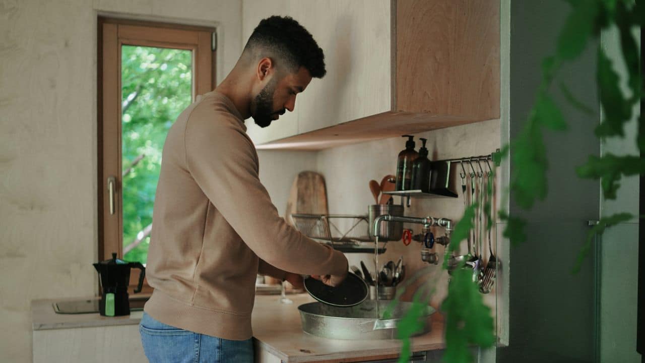A man with a beard is standing at a kitchen sink, washing a plate.
