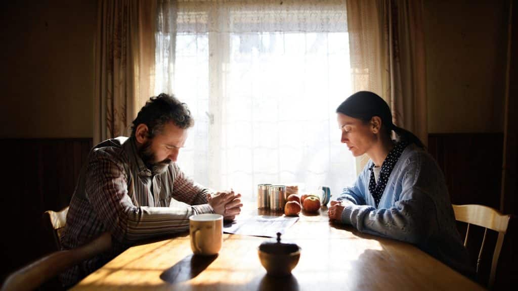 A man and a woman are sitting across a wooden table from each other, both looking down with their hands clasped.