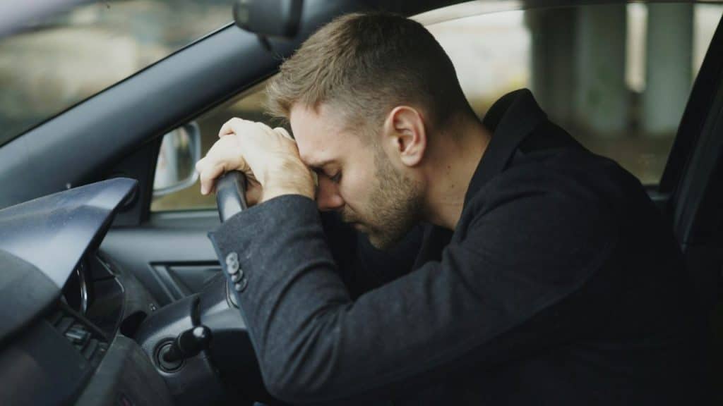 A distressed man leans his head onto the steering wheel of a car.