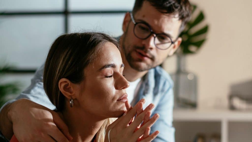 A man with glasses gently comforts a sad woman with her hands clasped.