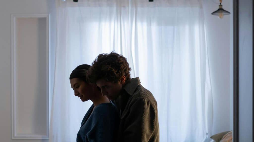 A couple standing in a softly lit bedroom, embracing quietly near the bed.