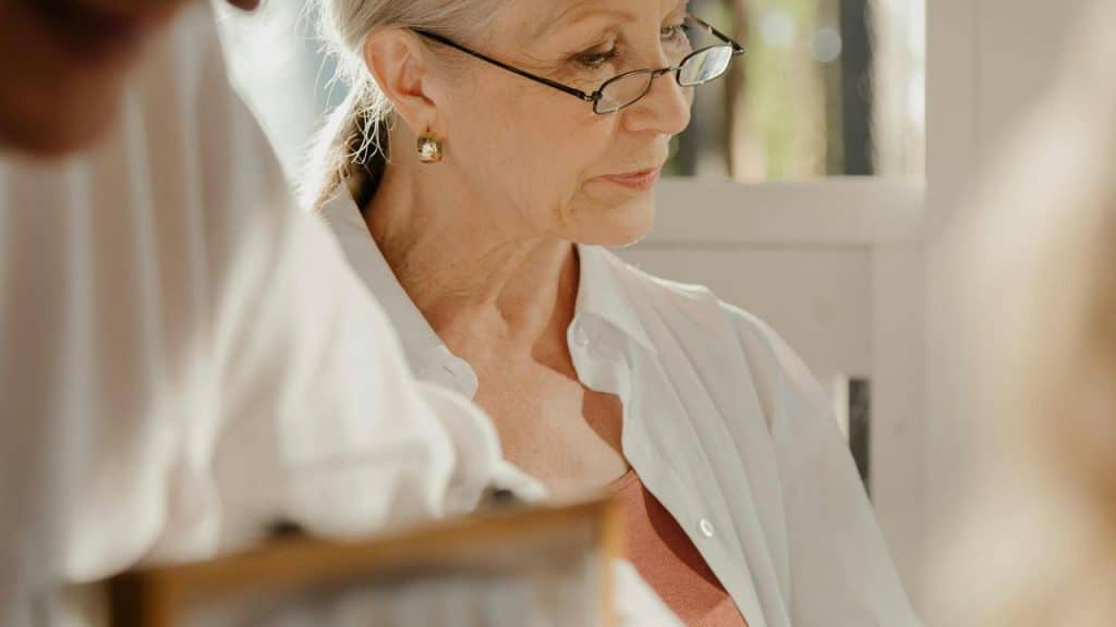 An elderly woman wearing glasses and a white shirt looking down thoughtfully.