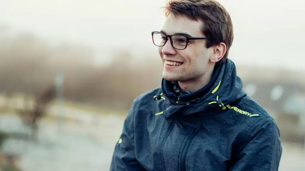 A man wearing glasses and a jacket smiling outdoors.