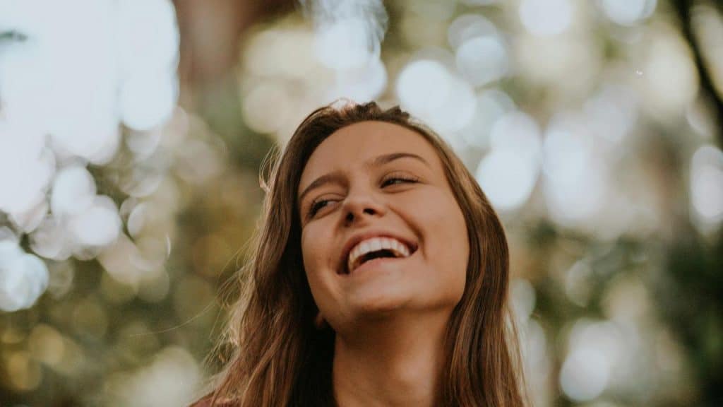 A woman laughing outdoors with soft, blurred greenery in the background.