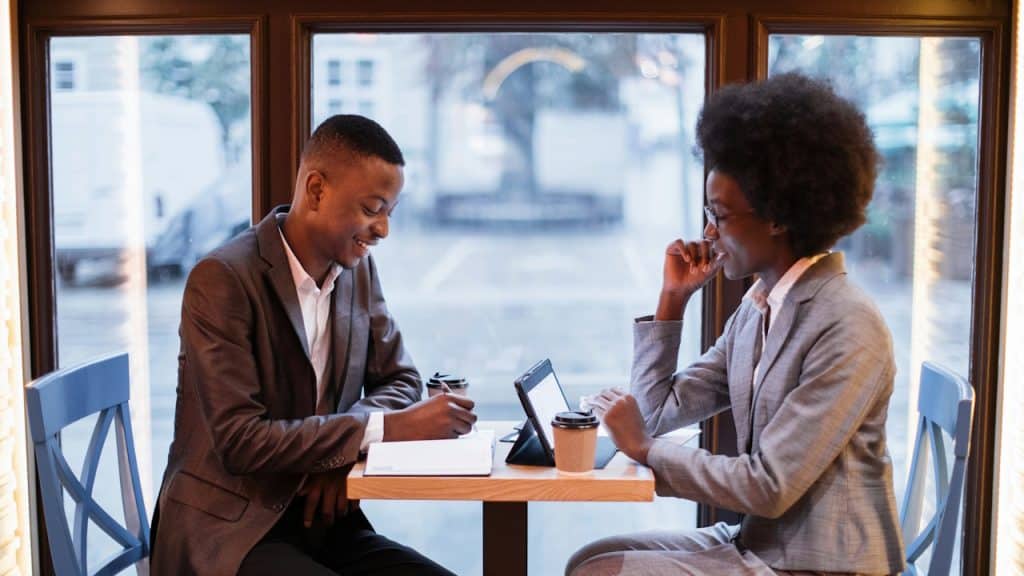 Two formally dressed people are sitting at a table in a cafe by a window, looking at a tablet and writing.