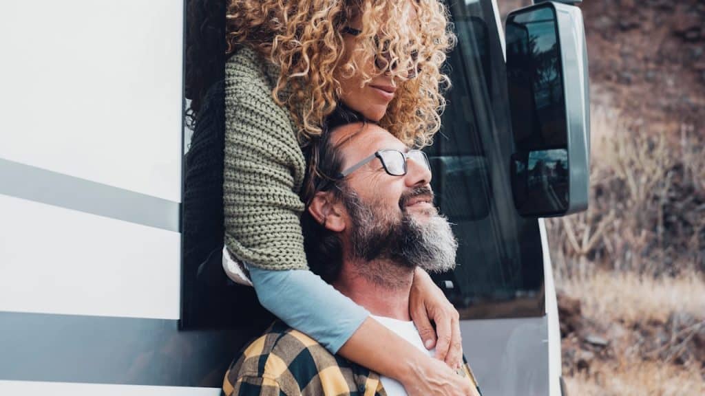 A woman leaning out of a wagon’s window and resting her hands on a bearded man’s chest, who’s leaning against the wagon.