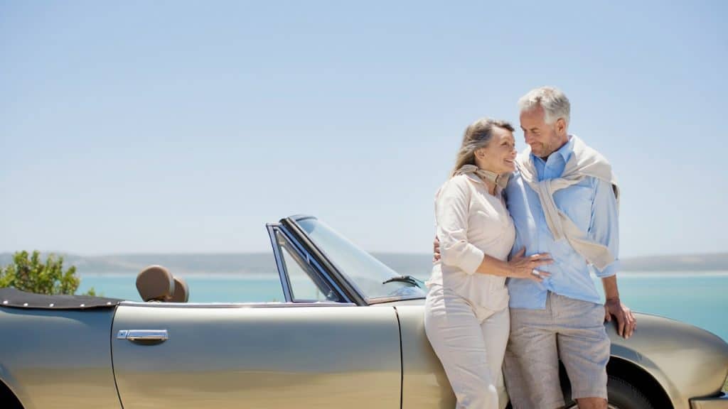 A couple standing close to each other while leaning against a convertible parked on a beach by the sea.