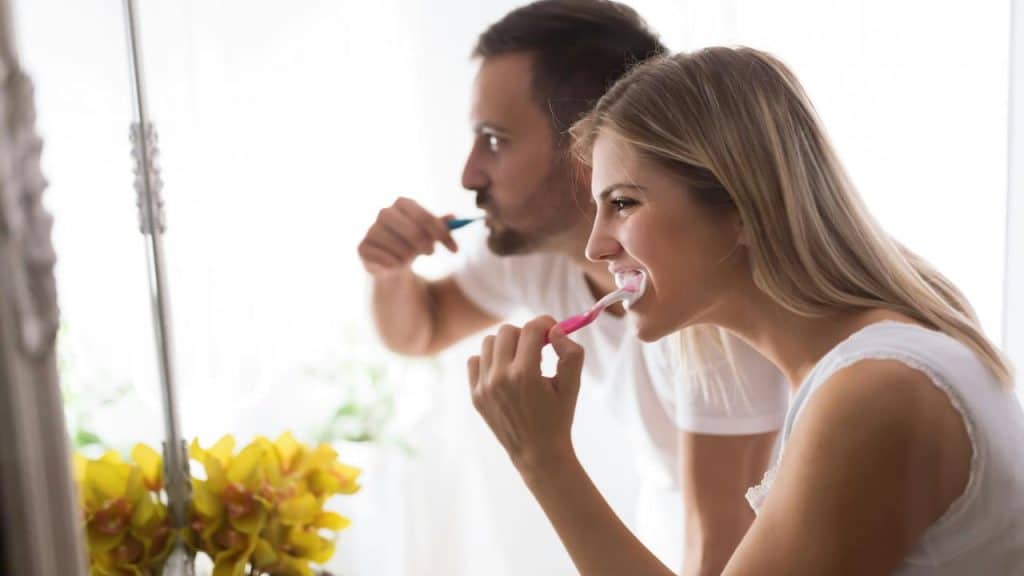 A man and woman brushing their teeth together in front of a mirror.
