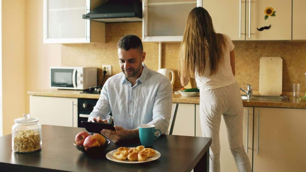 A man smiles while using a tablet at a kitchen table; a woman works at the counter behind him.