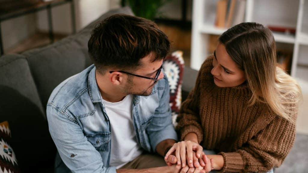 A man and woman sit close on a couch, holding hands and looking at each other.