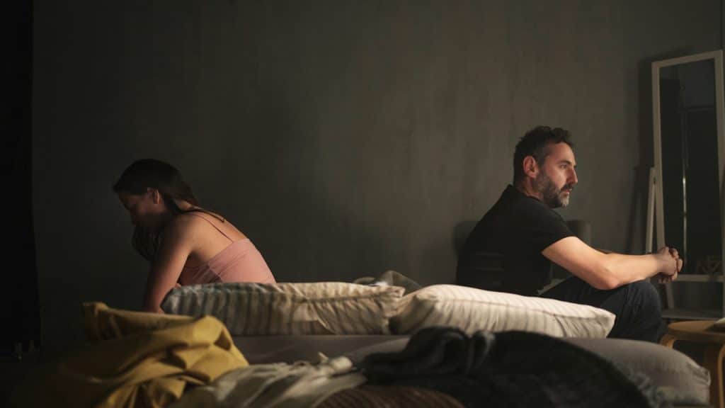 A man and a woman sit facing away from each other on a bed in a dimly lit room.
