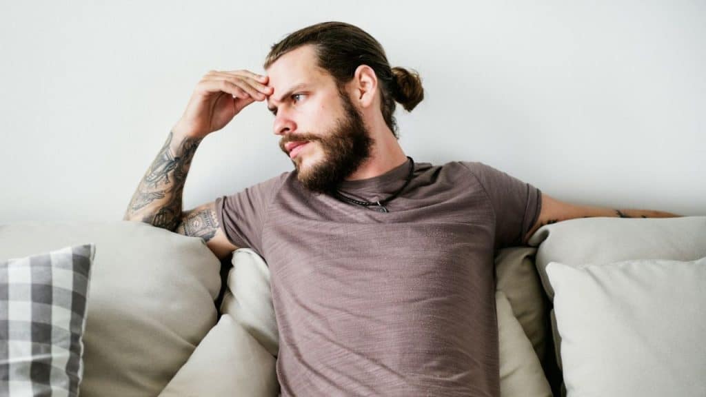 A bearded man with a top knot and tattooed arm is sitting on a couch, looking away.