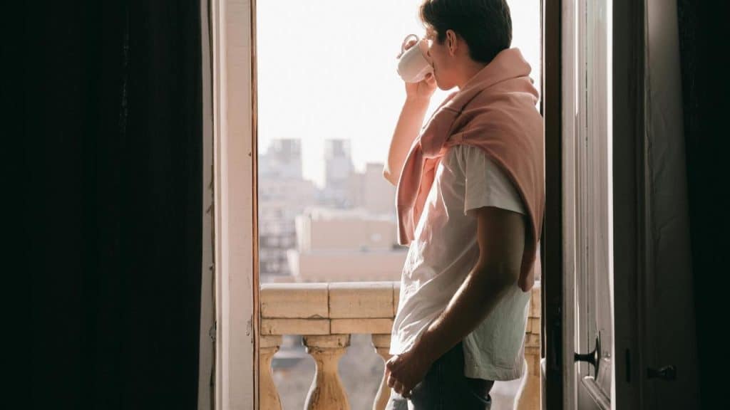 A man standing on a balcony drinking from a mug while looking at the city.
