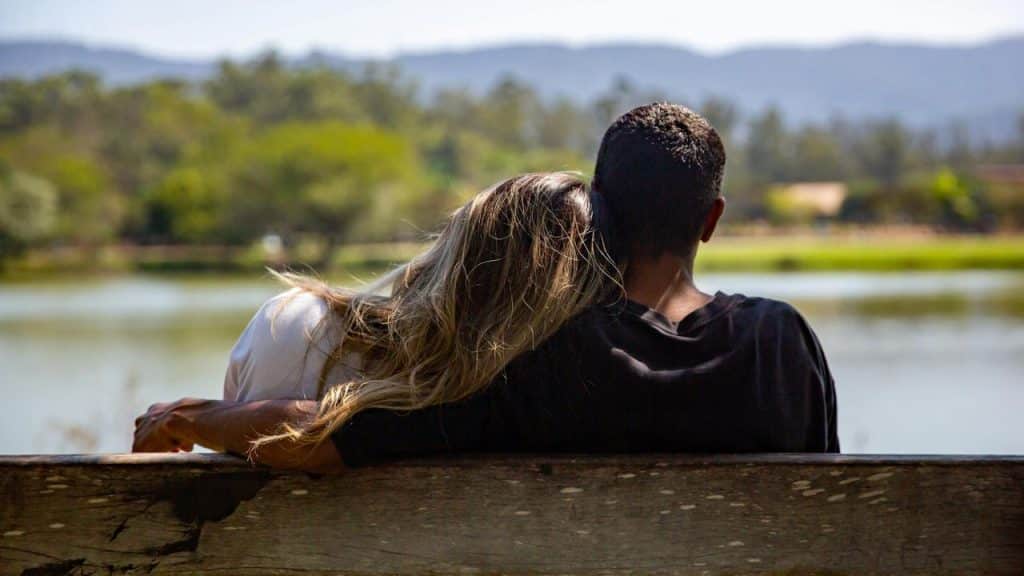 A couple sitting on a bench by the water with heads resting together.