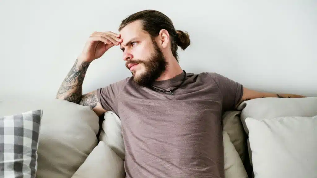 A bearded man with a top knot and tattooed arm is sitting on a couch, looking away.