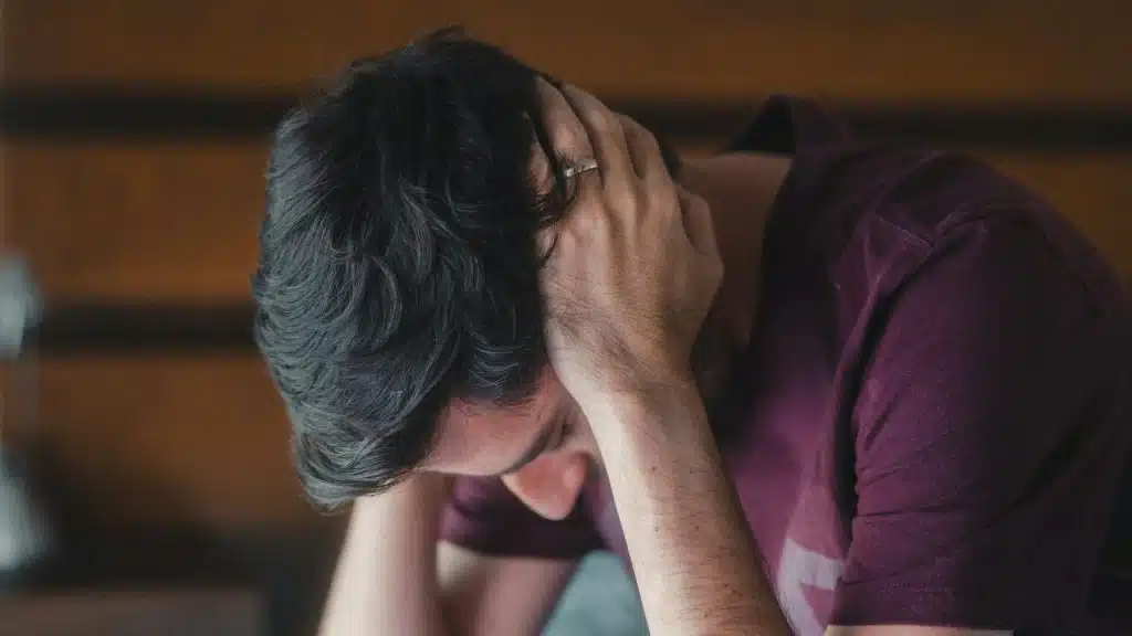 A man in a purple shirt sits with his head in his hands.