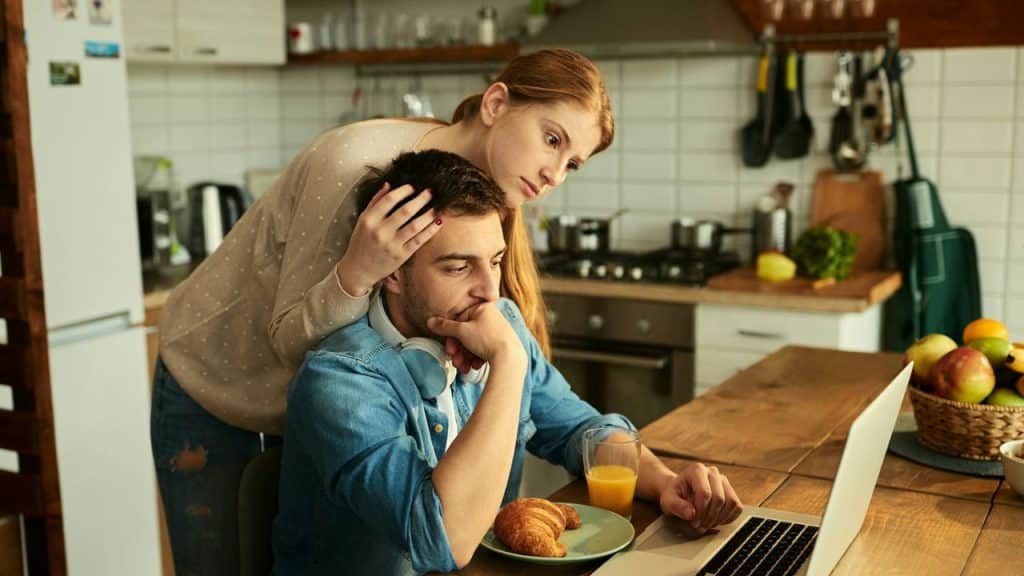 Woman comforting a stressed man looking at a laptop in a bright, modern kitchen.