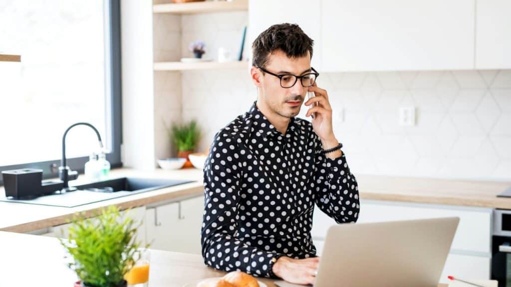 Man in a polka-dot shirt working on a laptop and phone in a kitchen.