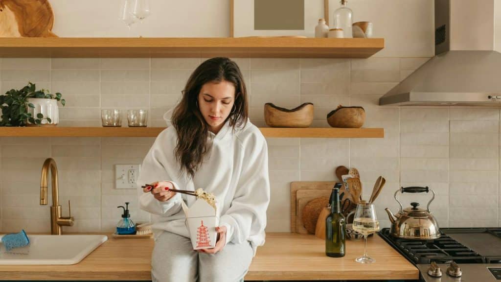 A woman sitting on a kitchen counter eating noodles from a takeout box.