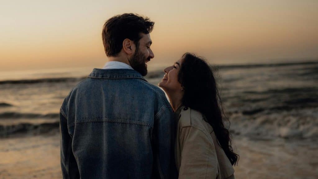 A couple smiling at each other while standing on a beach at sunset.
