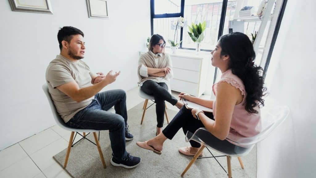 Three people sit in a brightly lit room with a window, engaged in a discussion.
