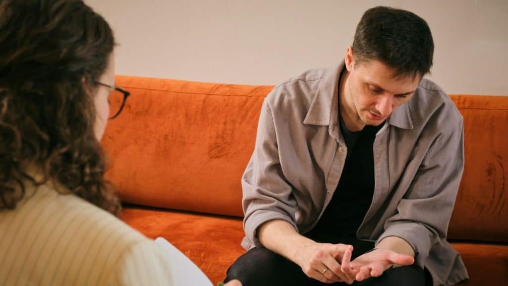 Man looking down and holding his hands on an orange couch, talking to a woman with glasses.
