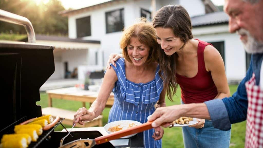 Two smiling women standing by a grill with a man, cooking food outdoors in a backyard setting.
