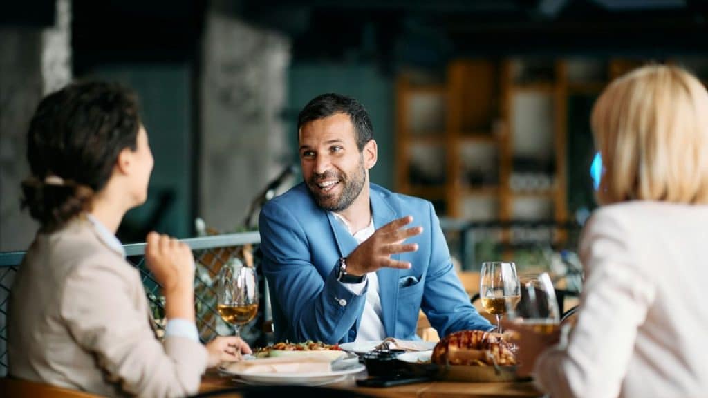 Smiling man in blue jacket talking with two women at a restaurant table.
